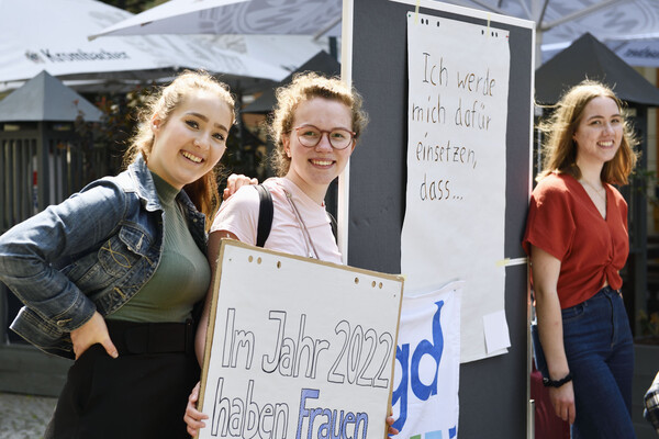 Drei Freiwillige werben für feministisches Engagement beim Aktionstag des FSJ Politik vor einer Stellwand