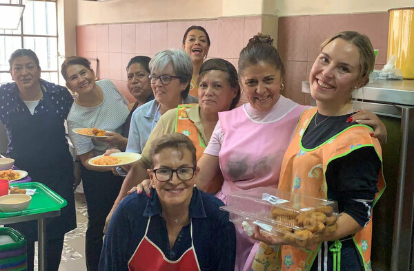 Eine Freiwillige und mehreren Frauen stehen in einer Küche mit Essen in der Hand und lächeln in die Kamera.
