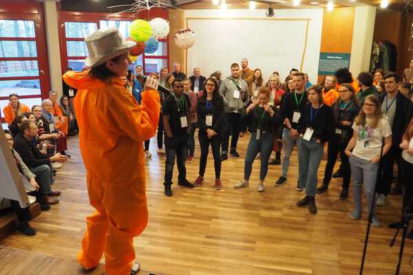 Eine Mitarbeiterin steht in einem orangenen Maleranzug vor Mitarbeiter*innen und Vereinsmitgliedern und spricht in ein Mikrofon.
